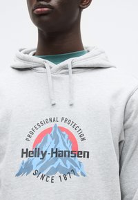 Γκρι φούτερ με κουκούλα που παρουσιάζει ένα γραφικό βουνού σε μπλε και κόκκινο, με το κείμενο «Επαγγελματική Προστασία» και «Helly-Hansen Από το 1877».