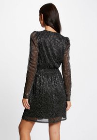 Zwarte jurk met doorzichtige lange mouwen, met een gestructureerde, metallic afwerking met zilveren draad. Getailleerde taille en uitlopende rok.