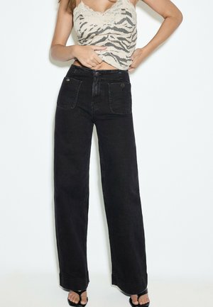 Person trägt hoch taillierte schwarze Jeans mit weitem Bein und Fronttaschen, ein Spitzenbesetztes Top mit Zebramuster und schwarze offene Sandalen.