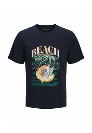 Garçon blond souriant, portant un T-shirt bleu marine avec l'inscription "BEACH" et un motif tropical, un short vert délavé jusqu'aux genoux, des chaussettes blanches et des baskets beige.