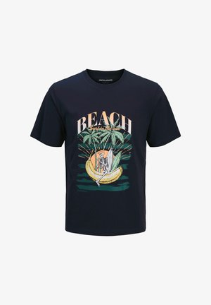 Blond jongetje glimlacht, draagt een marineblauw T-shirt met "BEACH" en tropische afbeelding, vervaagde groene knielange korte broek, witte sokken en beige sneakers.