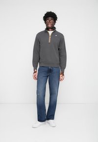 Felpa grigia con zip e accenti arancioni, abbinata a jeans blu e sneakers bianche. Tessuto morbido, vestibilità casual e polsini a costine.