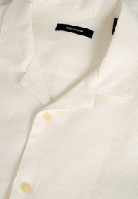 Camisa de lino crema con cuello, dos botones amarillos y una textura suave. La etiqueta dice "Matinique."