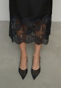 Abito in satin nero con orlo in pizzo floreale che mostra motivi intricati. Abbinato a décolleté slingback nere con punta a punta. Texture liscia.