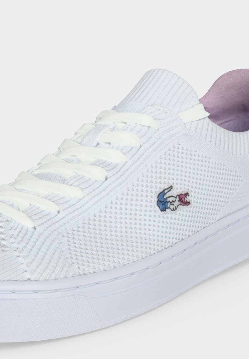 Weißer Stricksneaker mit weißen Schnürsenkeln, einer strukturierten Oberseite und einem kleinen blau-pinken bestickten Logo an der Seite.