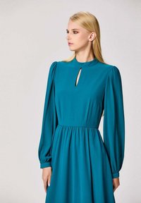 Robe longue à manches longues en bleu sarcelle avec un col haut et une découpe en forme de trou de serrure. Présente une taille froncée et une jupe fluide en tissu texturé. Design minimaliste.
