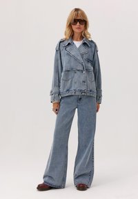 Kobieta w oversize'owej dżinsowej kurtce i szerokich spodniach dżinsowych, w kolorowych okularach przeciwsłonecznych i brązowych butach, stojąca na tle jasnej ściany.