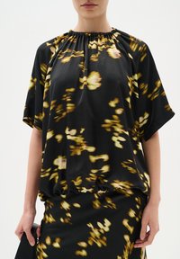Blusa nera con motivi floreali gialli, maniche corte, scollo arricciato e orlo elastico. Il tessuto liscio cade in modo morbido.