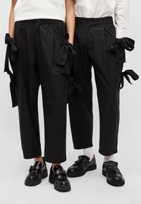 Calças pretas com grandes bolsos laterais, apresentando laços, um corte descontraído e uma textura suave. Combinadas com sapatos pretos robustos.