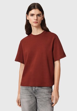 Jeune femme aux longs cheveux bruns portant un t-shirt marron uni à manches courtes et un jean gris clair, debout devant un fond neutre.