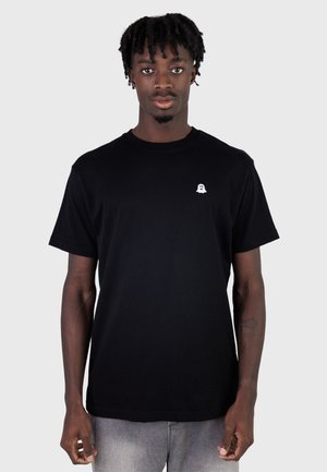 Jeune homme aux dreadlocks courtes portant un t-shirt noir avec un petit logo de fantôme blanc sur le côté gauche de la poitrine, debout devant un fond clair.