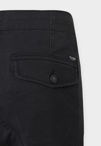 Mustat kangashousut, joissa takatasku nappikiinnityksellä ja pieni merkkilappu, kirjailtua yksityiskohtaa ja sileä pinta.