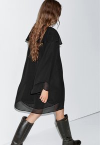 Schwarzes Shiftkleid mit einem geschichteten Rüschenausschnitt und durchsichtigen Ärmeln, kombiniert mit knielangen, dunkelbraunen Lederstiefeln.