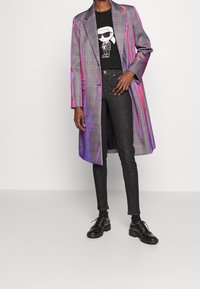 Rutig, skimrande lång kappa i grått och lila; svart grafisk t-shirt; mörka skinny jeans; svarta klumpiga skor med nitar.