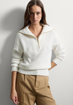 Pullover bianco lavorato a maglia con colletto zip, caratterizzato da una trama a costine e vestibilità comoda. Abbinato a pantaloni color beige.