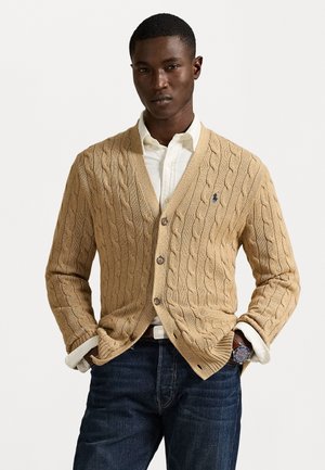 Beige cable-knit cardigan with a V-neck, wooden buttons, and a small logo. Paired with a white shirt and dark denim jeans.