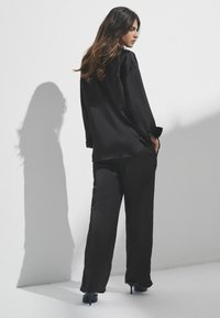 Kvindelig i sort løsbluse og bukser iført sorte høje hæle, stående mod en hvid væg med skygge kastet bag sig.