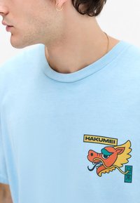 Light blue cotton t-shirt featuring a colorful dragon graphic and text “HAKUMEI” on the left chest. Ribbed crew neckline, short sleeves.