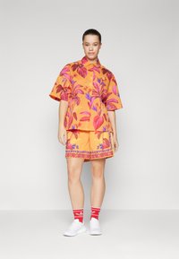 Oransje blomstret skjorte og shorts sett med store rosa og lilla bladmønstre, korte ermer og dekorativ kant på shortsene. Hvite sneakers.