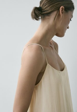 Femme avec les cheveux attachés en chignon, portant une robe sans manches beige clair, regardant vers le bas et sur le côté sur un fond gris clair uni.