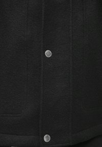 Sort uldfrakke med tekstureret overflade, trykknaplukning og diskrete syninger langs forsiden og kanten.