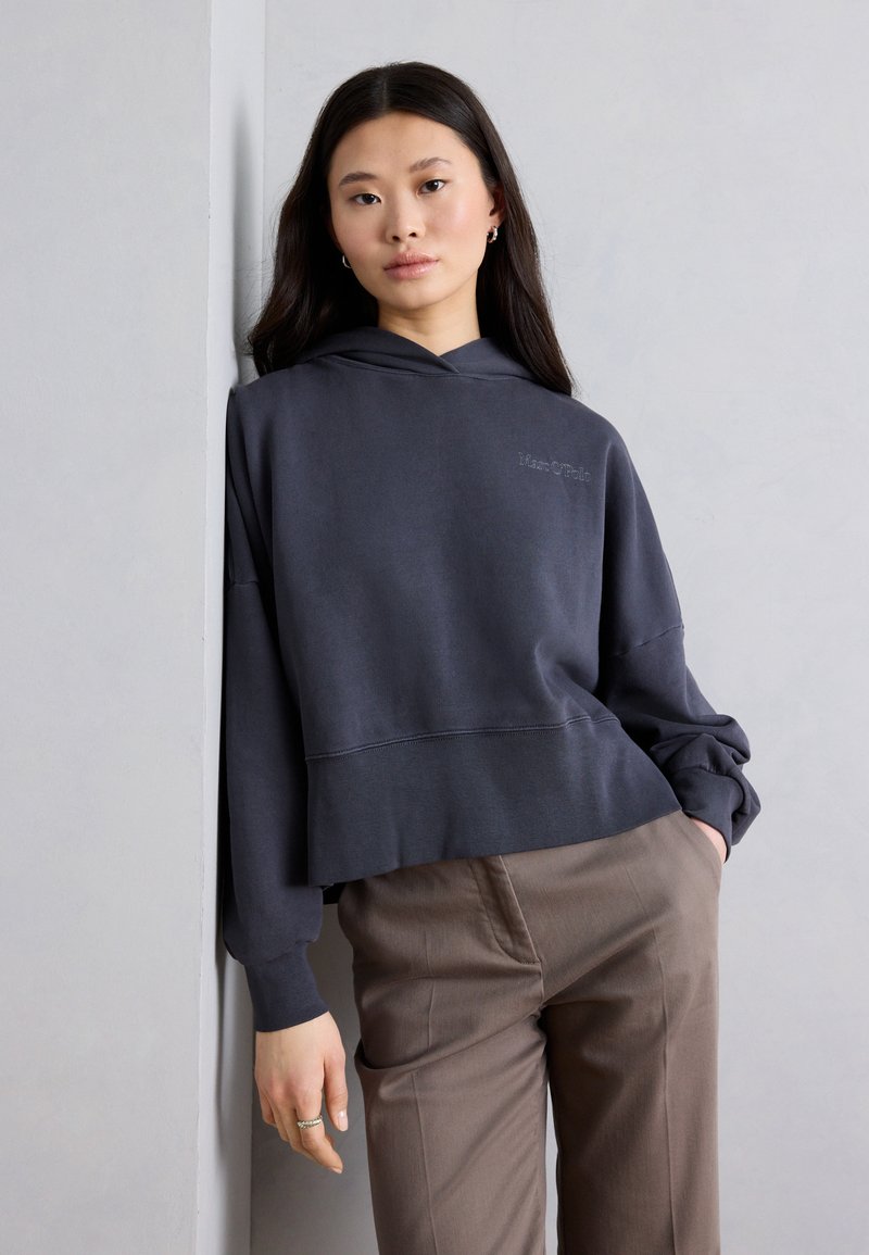 Νεαρή γυναίκα με μακριά σκούρα μαλλιά, ντυμένη με σκούρο γκρι κοντό φούτερ με κουκούλα και καφέ παντελόνια, στηριζόμενη σε έναν ανοιχτό γκρι τοίχο.