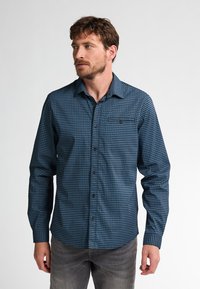 Camicia a maniche lunghe blu con motivo, colletto con bottoni, dotata di taschino sul petto e tessuto testurizzato. Jeans grigi visibili sotto.