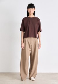 Donna in piedi che indossa una maglietta corta oversize marrone, pantaloni beige larghi e scarpe slip-on bianche su uno sfondo chiaro e semplice.