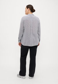 Chemise rayée à manches longues en noir et blanc, col boutonné, coupe relaxée, associée à un pantalon sombre et des chaussures claires.