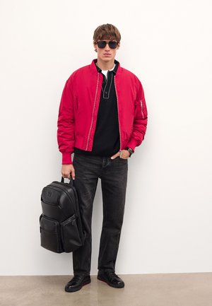 Giacca bomber rossa sopra una maglietta nera, jeans neri, sneakers nere e uno zaino nero con più tasche. Gli occhiali da sole completano il look.