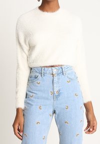 Pull en maille blanche duveteuse avec un col rond, associé à un jean bleu clair orné d'une broderie arc-en-ciel. Texture douce, coupe courte.