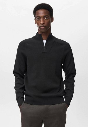 Pull en tricot noir avec un col zippé à mi-hauteur, présentant une finition texturée et des poignets ajustés, porté par-dessus un t-shirt blanc. Fond uni.