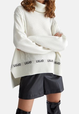 Witte gebreide coltrui met kabelgebreide accenten en een logoband aan de zoom, gecombineerd met zwarte leren shorts en glanzende laarzen.