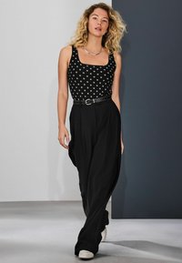 Black polka dot tank top with a square neckline, paired with loose black trousers and white sneakers, featuring a black belt with a metallic buckle.