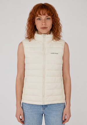 Femme aux cheveux roux bouclés portant un gilet matelassé sans manches blanc cassé zippé, associé à un jean bleu clair, face à l'avant.