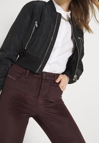 Černá bomber bunda se zipy, kontrastující s bílou košilí s knoflíky. Bordó džíny s mírným leskem a předními kapsami.