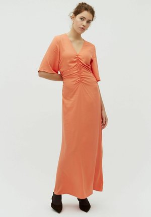 Młoda kobieta stojąca z jedną ręką za plecami, ubrana w długą, marszczoną koralową suknię i czarne szpiczaste buty na jednolitym tle.