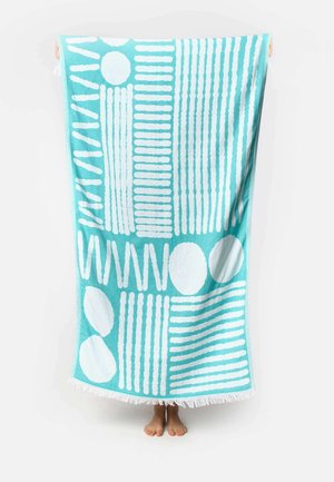 Personne debout pieds nus derrière une serviette turquoise avec des motifs géométriques blancs et des bords à franges.