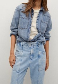 Chemise en denim bleu clair avec deux poches poitrine, boutonnée sur un haut rayé blanc. Pantalon en denim bleu clair taille haute avec des coutures visibles.