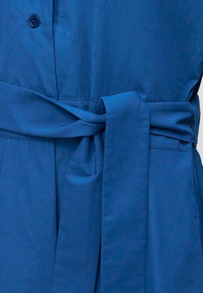Μπλε υφασμάτινο ένδυμα με ζώνη μέσης δεμένη σε κόμπο κάτω από ένα μόνο ορατό κουμπί στο μπροστινό μέρος.