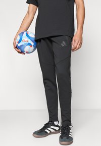 Μαύρο αθλητικό παντελόνι με στενή σχεδίαση, με υφή υλικού, πλαϊνές τσέπες και λογότυπο Adidas. Μια μπλε και κόκκινη μπάλα ποδοσφαίρου κρατιέται στο αριστερό χέρι.