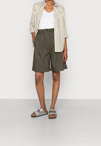 Stribet beige og hvid skjorte med knapper, hvid v-hals top, olivengrønne shorts og sølvfarvede sandaler med korksåler, stående på en hvid baggrund.
