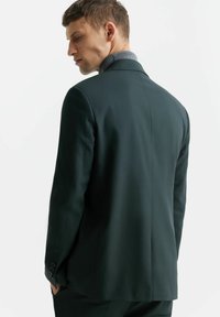 Ciemnozielona marynarka z szerokimi ramionami, zapinana na jeden guzik, z klapami. Posiada gładką fakturę materiału i elegancki krój.