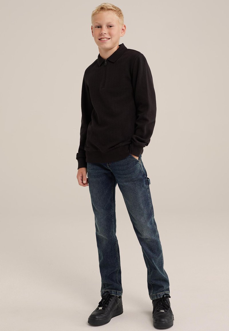 Schwarzes Langarm-Polo mit Reißverschlusskragen, kombiniert mit blauen Jeans und schwarzen Turnschuhen. Der Stoff hat eine strukturierte Oberfläche.