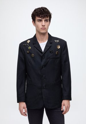 Un joven posa sobre un fondo liso y claro, con una chaqueta de traje negra adornada con diversos broches sobre una camisa blanca.