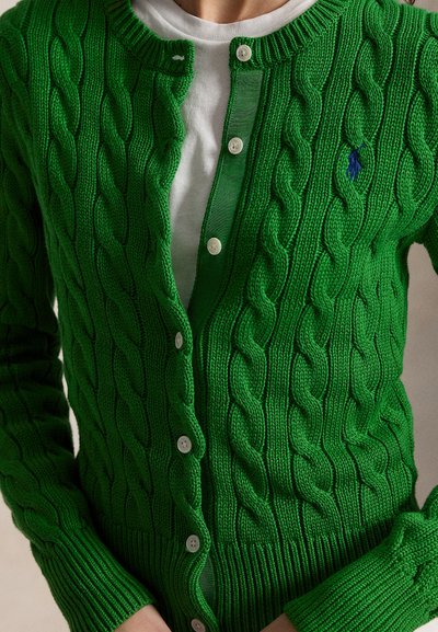 Zelený kardigán s pleteným vzorom a bielymi gombíkmi, nosený cez biele tričko s okrúhlym výstrihom, s malým modrým vyšívaným logom na hrudi.