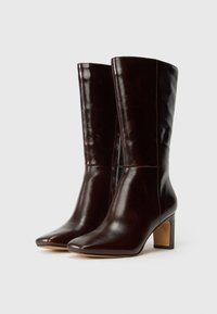Par de botas de cuero de medianoche marrón oscuro, hasta la mitad de la pantorrilla, con punteras puntiagudas y tacones en bloque, exhibidas sobre un fondo claro y liso.