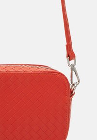 Červená tkaná kožená crossbody taška se stříbrným zipem a odnímatelným popruhem na bílém pozadí.