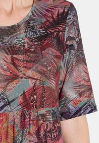 Nahaufnahme einer Person, die ein farbenfrohes, gemustertes Kurzarmoberteil mit abstrakten roten, lila und grauen Blumen- und Blattdesigns trägt.