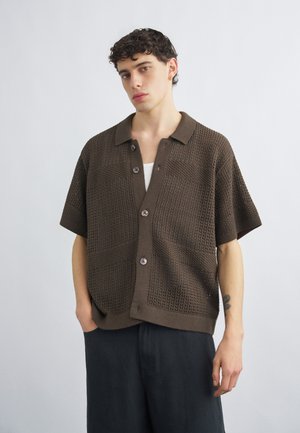 Νέος άνδρας με κοντά σγουρά μαλλιά που φοράει καφέ πουκάμισο με κοντά μανίκια πλεγμένο ανοιχτό, με κουμπιά, πάνω από λευκό φανελάκι και σκούρο παντελόνι.